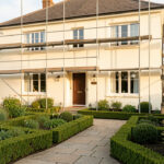 Rénovation de façade maison : étapes, matériaux et contrôles Maison rénovée à la façade claire, entourée d’un jardin soigneusement entretenu, avec un échafaudage discret et éclairée par une lumière naturelle.